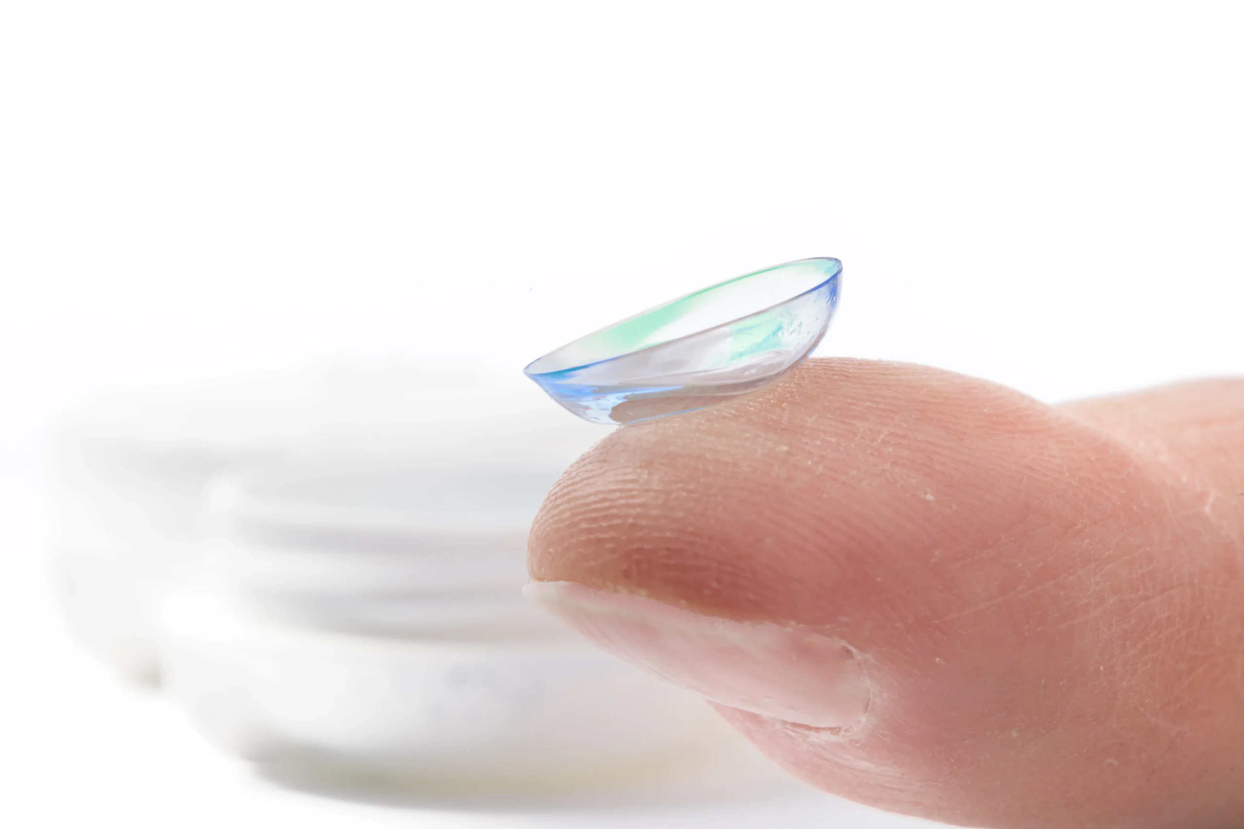 Primer plano de un lente de contacto graduado blando con tonos azules y verdes, perfectamente curvado sobre el pulpejo de un dedo.