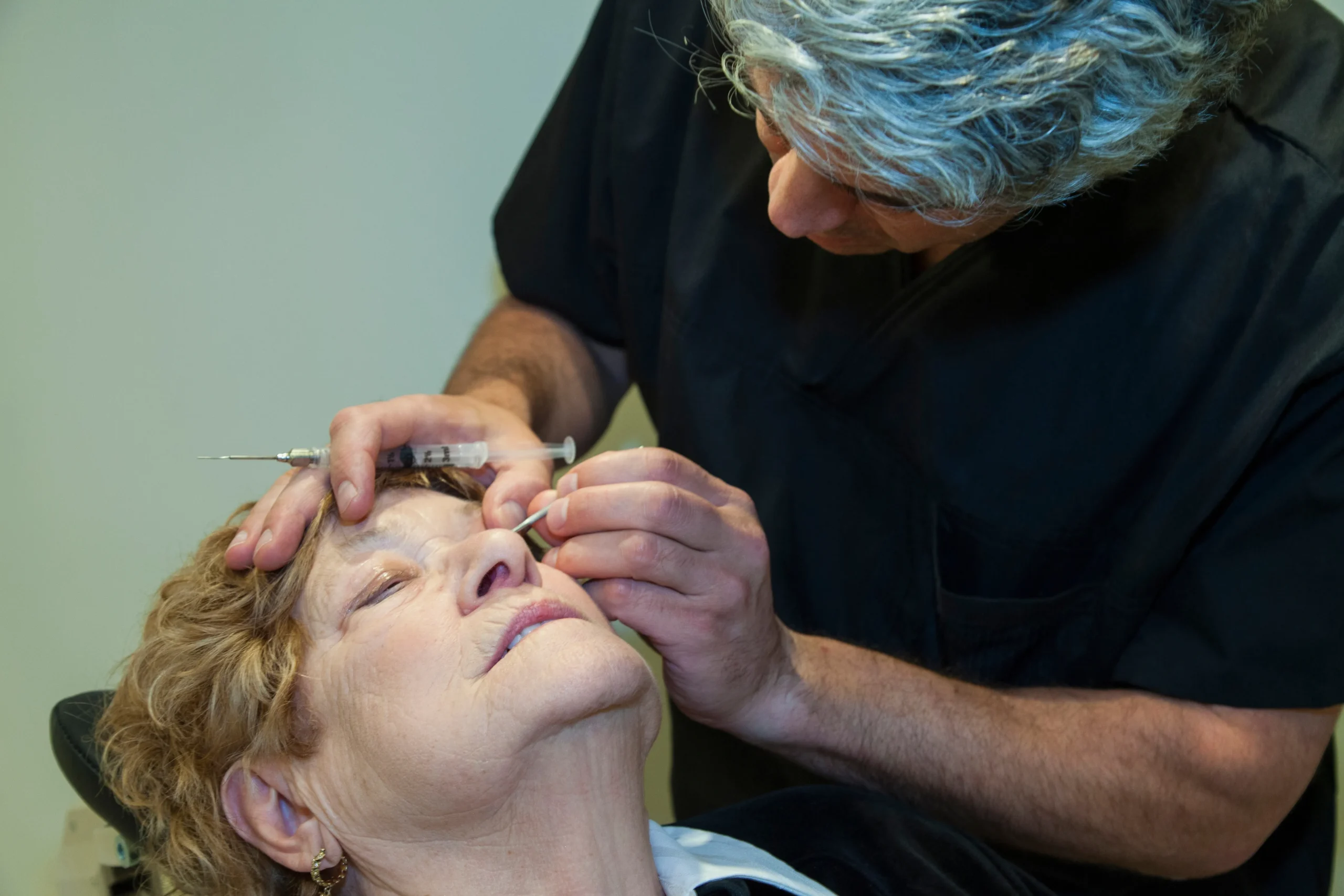 Profesional de la salud inyectando ácido hialurónico cerca del área de los ojos de la paciente en una consulta de medicina estética pre-blefaroplastia