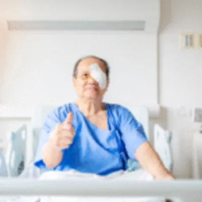 A patient in a blue gown is sitting up, smiling broadly and giving a thumbs-up after successfully completing his eye surgery in Mexico.