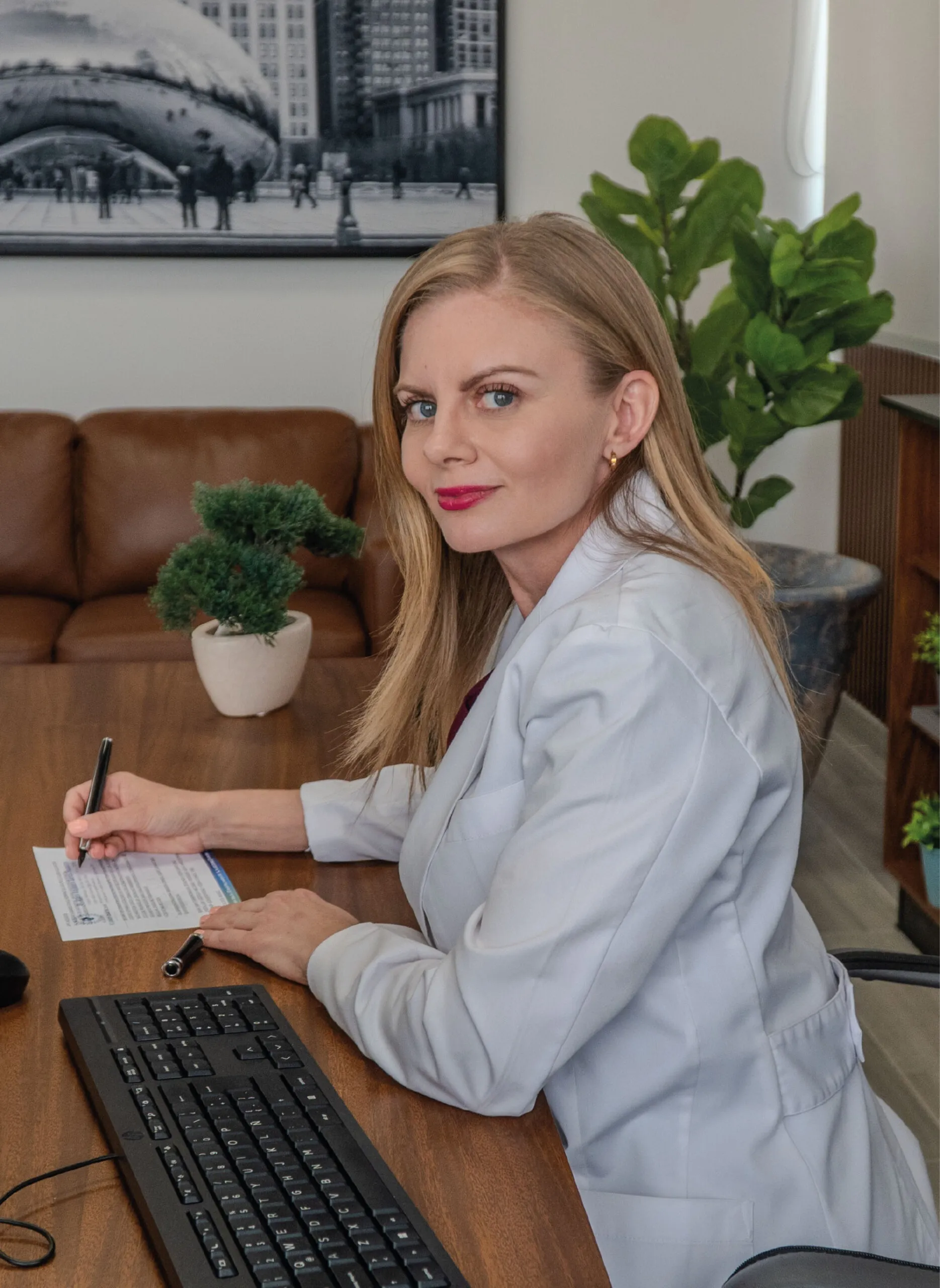 A blonde female ophtlamologist in Mexico in a white coat sits at a wooden desk with a bonsai tree and writes on paper.