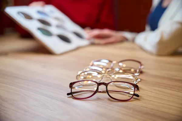 Close up of various fashion eyeglasses and ophthalmic lenses with a person holding a lens sample book in the background