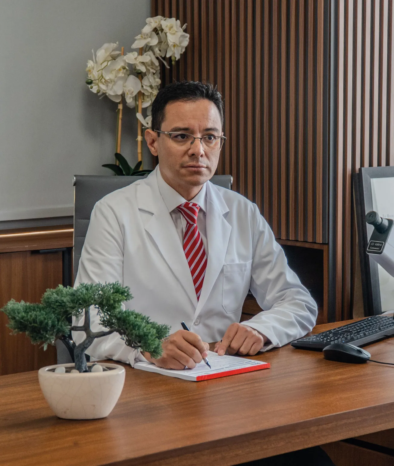 A male ophtlamologist in Mexico wearing a white coat, glasses, and a red striped tie writes on a pad at a desk.