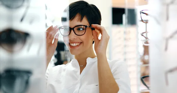 A happy young woman testing a pair of stylish glasses with ophthalmic lenses while looking in the mirror at the boutique